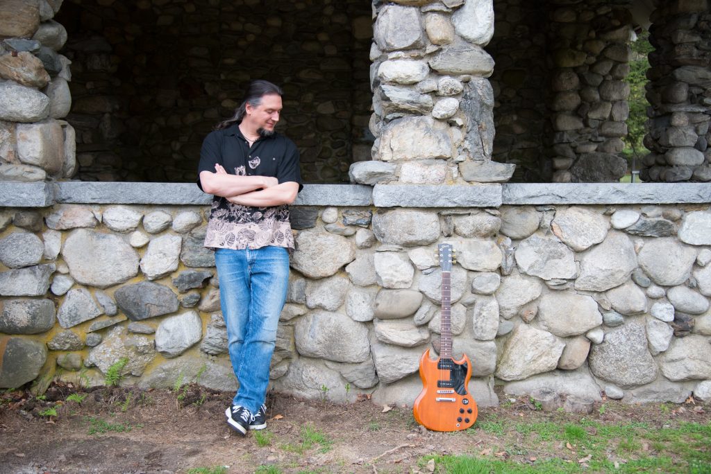 Mike Loce looking at a brown electric guitar in front of a stone structure