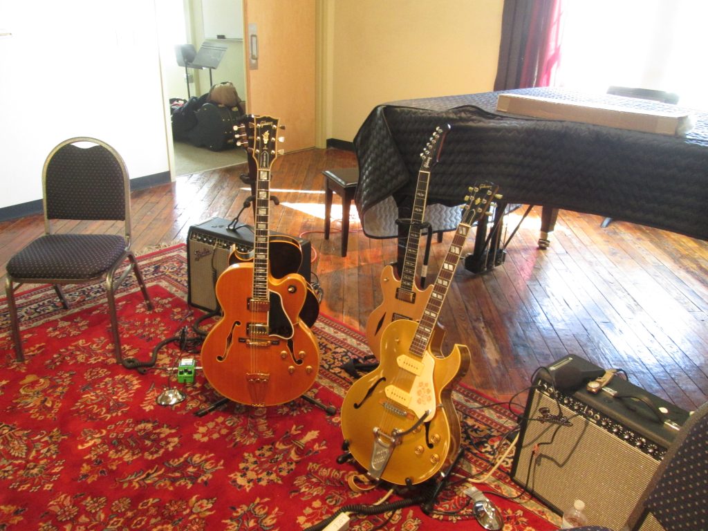 Three electric guitars and two amps on a red carpet in a recital hall